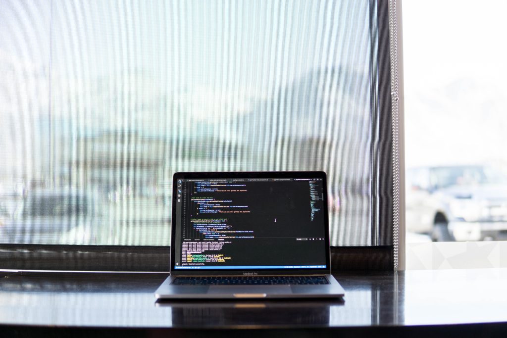 A laptop computer with code on the screen sitting on a desk in front of a window